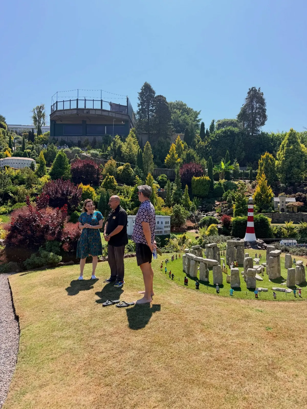 Three people standing on the lawn beside a miniature Stonehenge at Babbacombe Model Village, a red and white lighthouse and tiny buildings on the hillside behind.