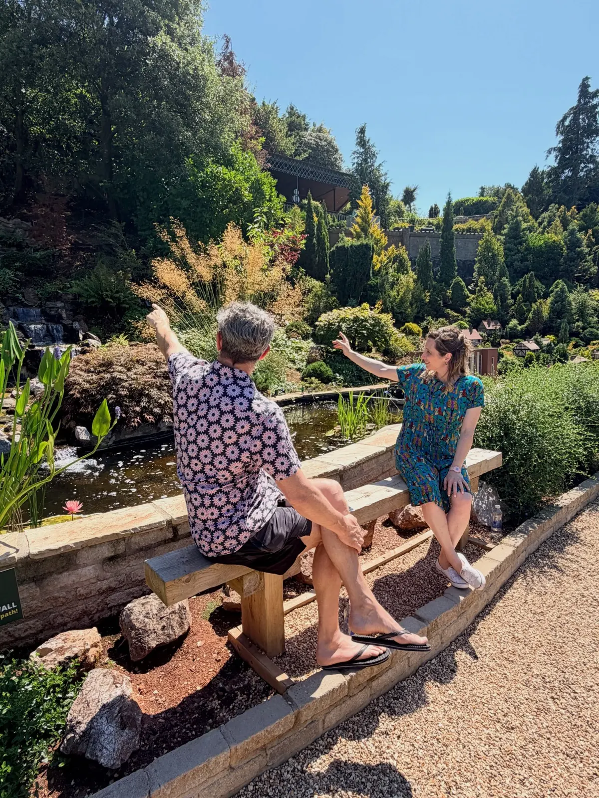 Isy Suttie and Dan Skinner on a bench beside a lily pond at Babbacombe Model Village, both pointing off at something in the miniature landscape.