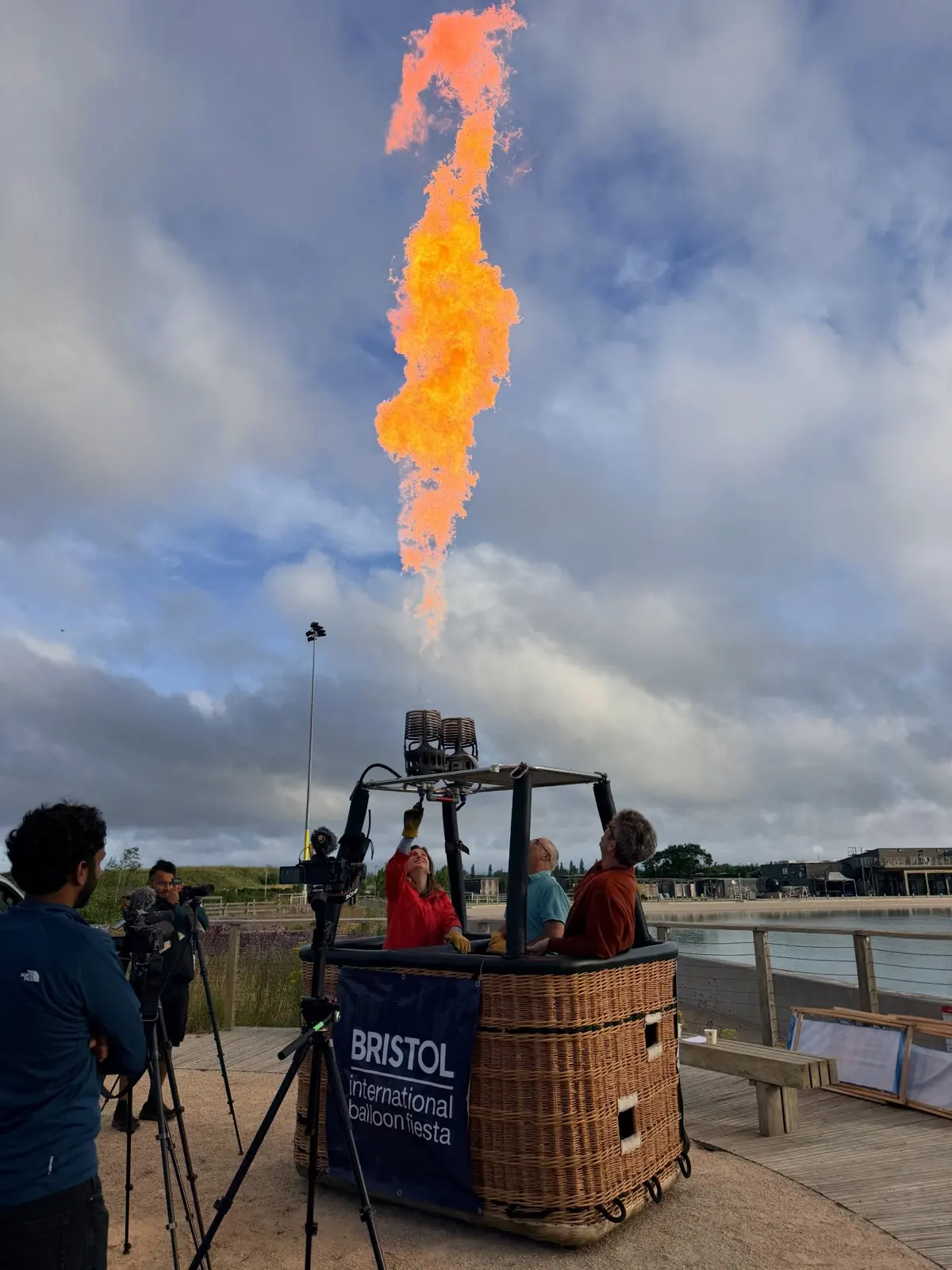 A burner flame roaring above a wicker basket stencilled Bristol International Balloon Fiesta, a camera operator filming from beside it.