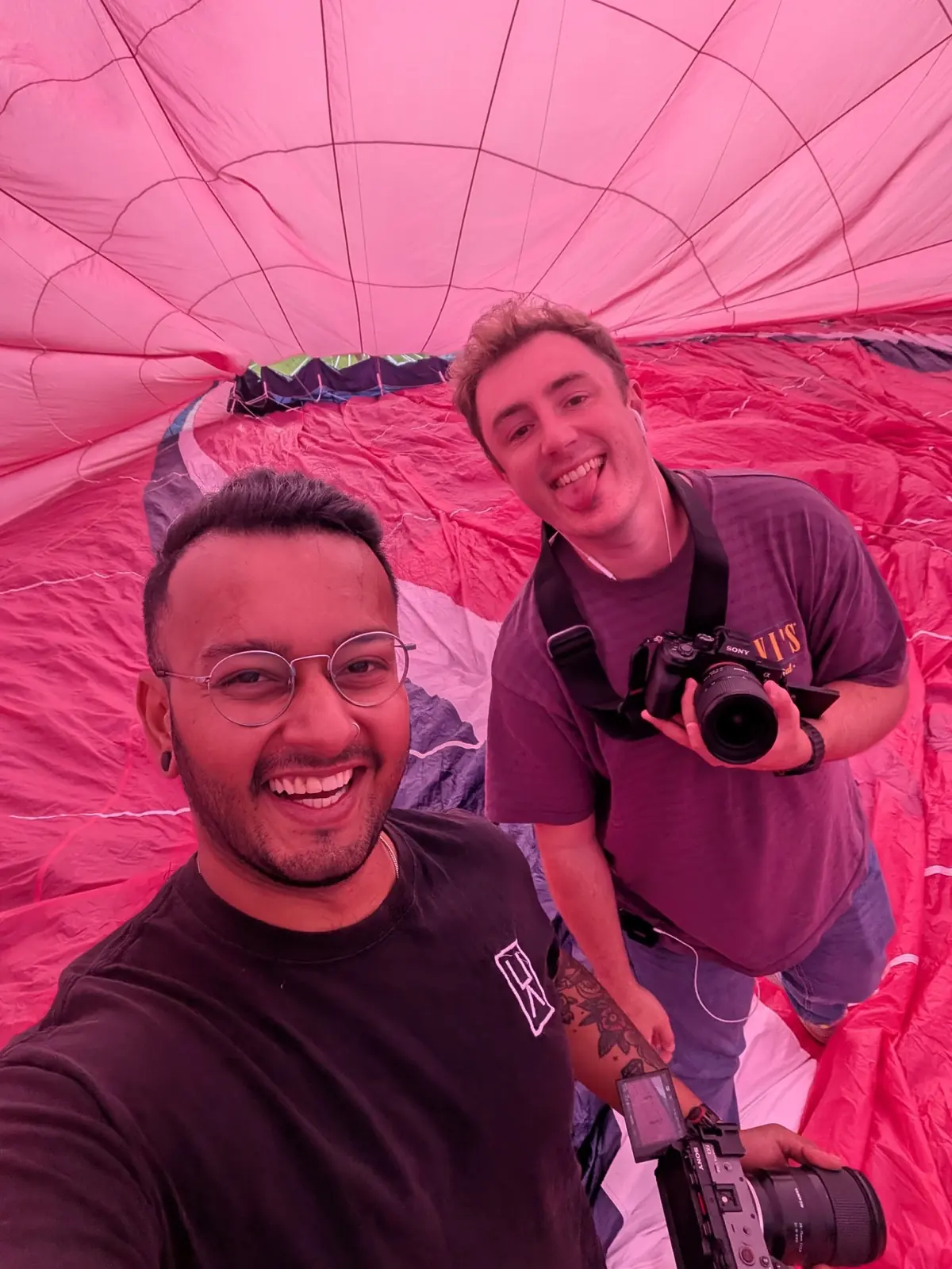 Two crew grinning for a selfie inside the pink balloon envelope, one holding a Sony camera, everything lit rose by the fabric.