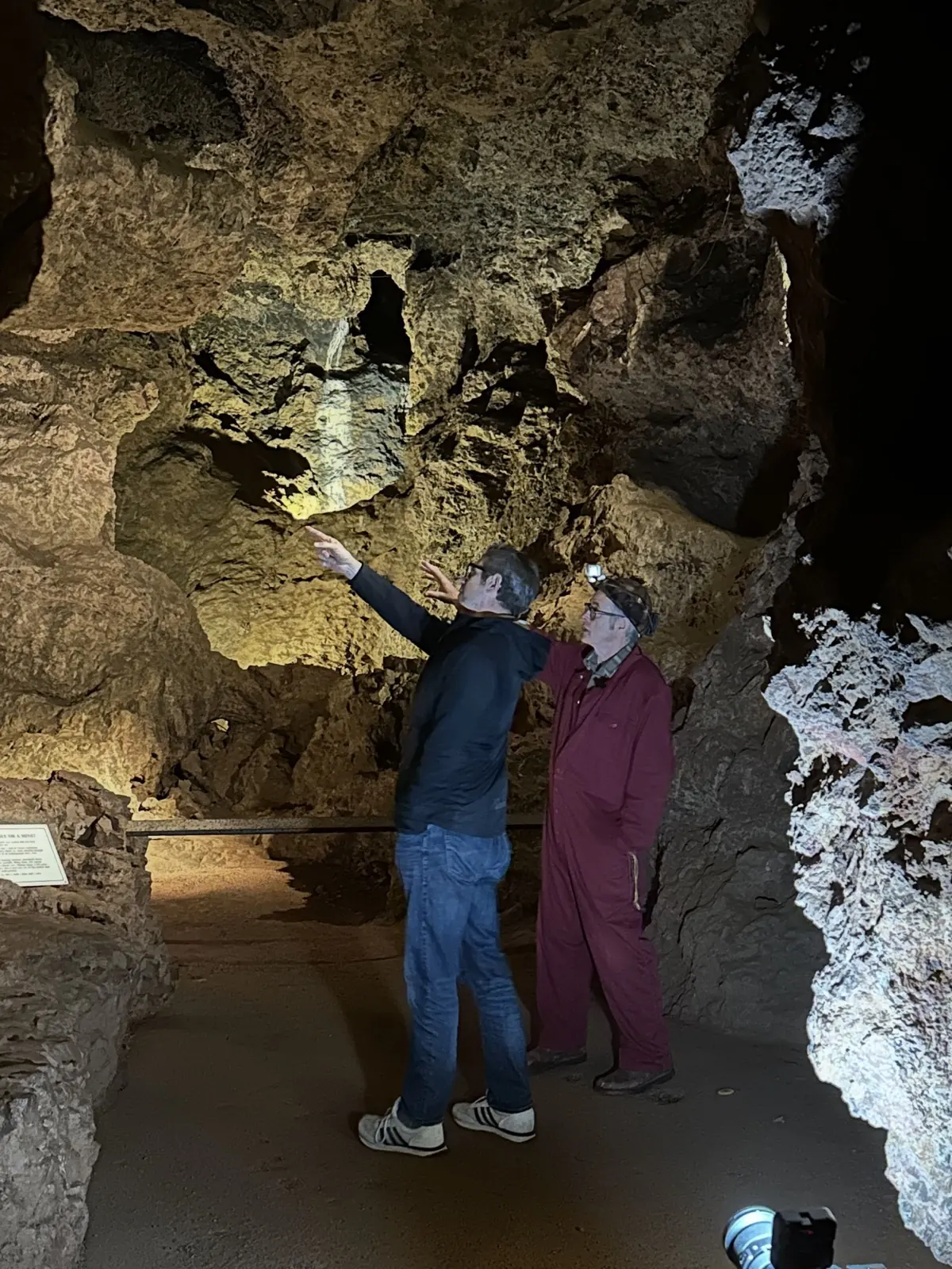Underground at Clearwell Caves: a freeminer in a maroon boilersuit and head torch points up at the rock ceiling while a second man looks where he is pointing.