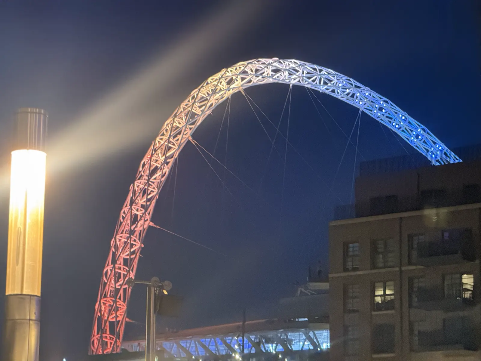 Wembley's arch lit pink fading to blue against the night sky, rising behind a block of flats and a glowing lamp post.
