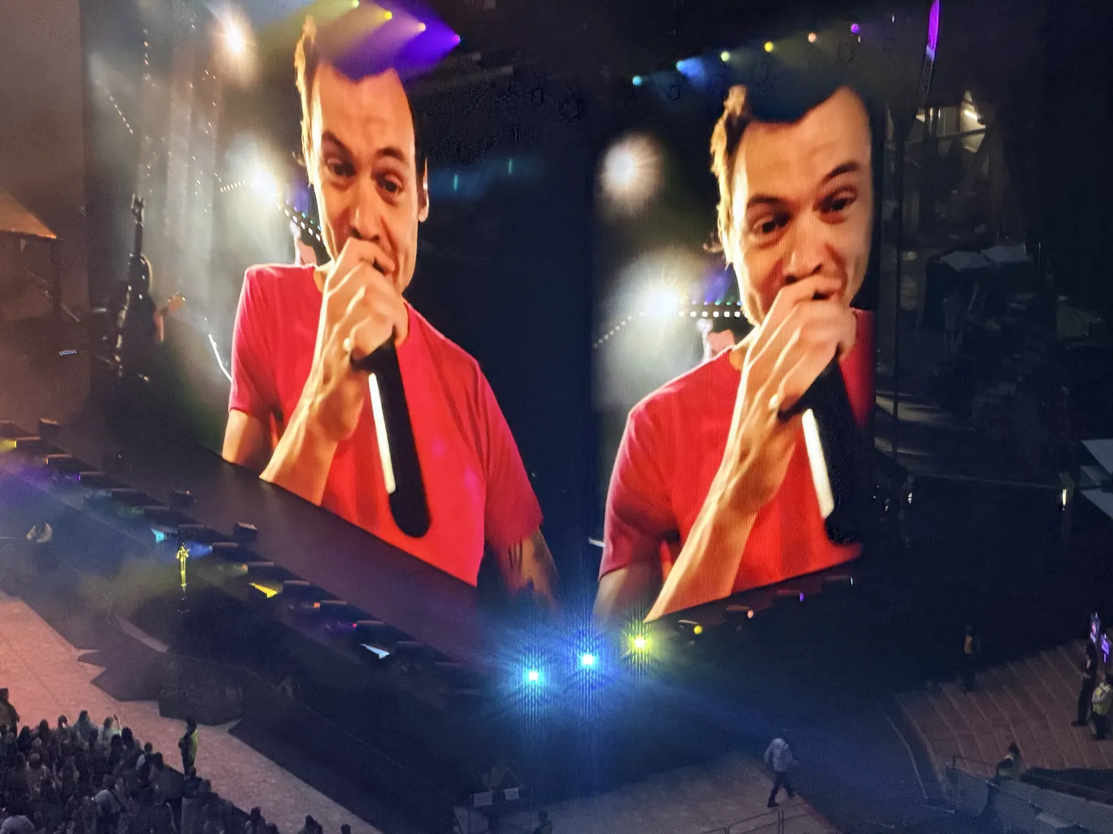 Harry Styles in a red T-shirt grinning into a microphone, shown twice across the giant stadium screens, stage lights flaring below.