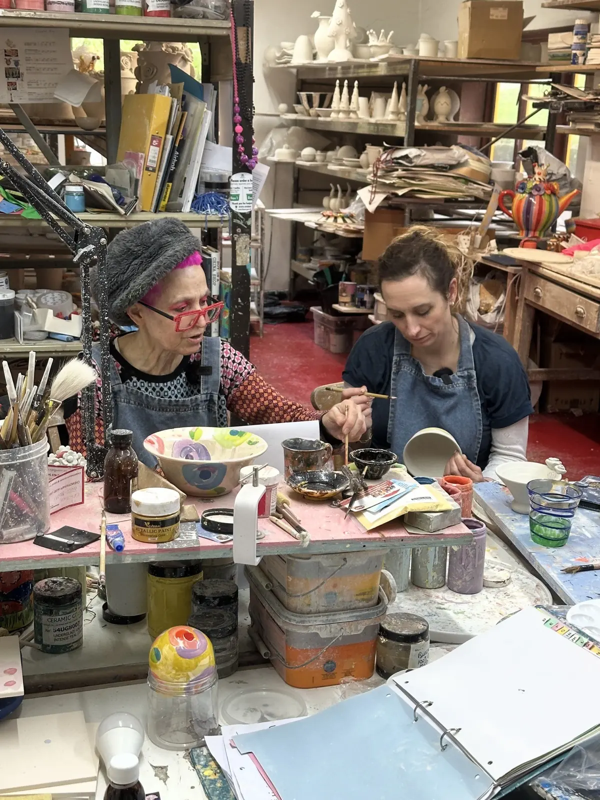 Mary Rose-Young, in pink hair and red glasses, painting pottery alongside Isy Suttie in a studio crowded with brightly coloured glazes and finished pieces.