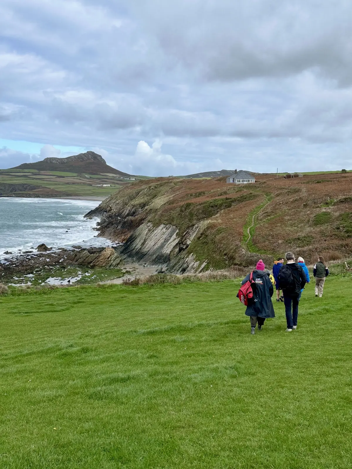 A line of people in coats and dryrobes walking a clifftop path down towards the sea in Pembrokeshire, under a pale grey sky.