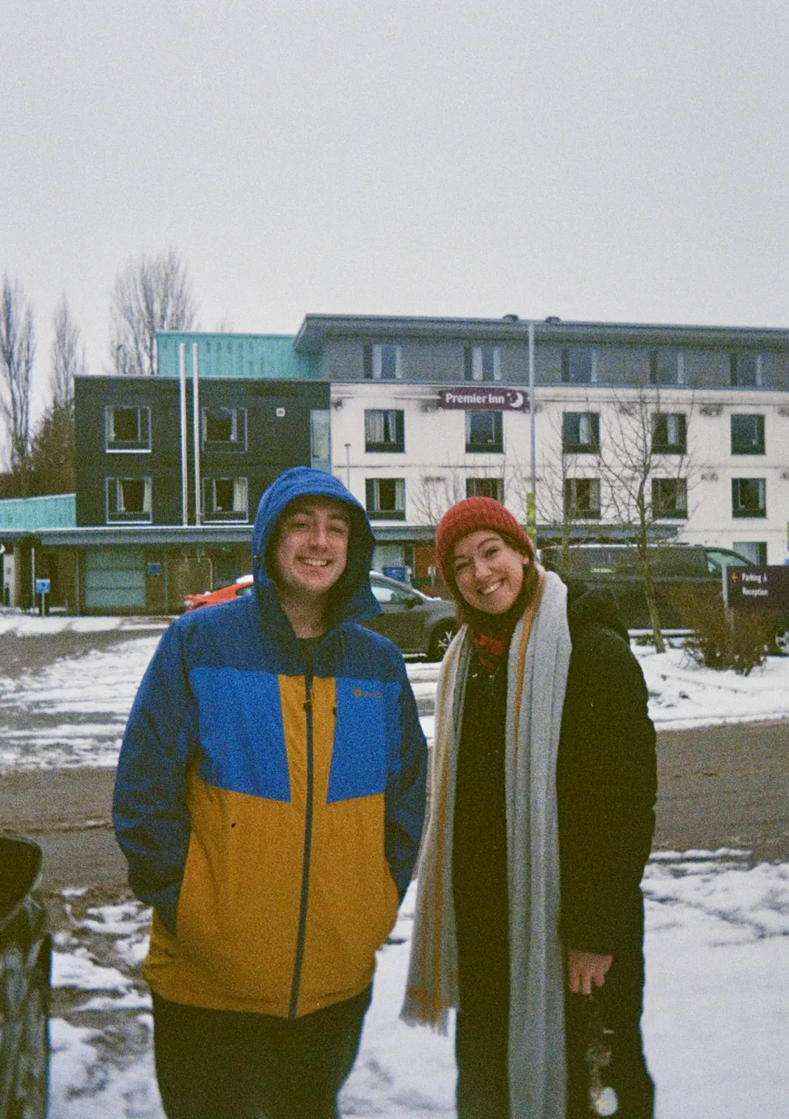 Adam Stoner in a blue and yellow hooded jacket and Sarah MacGillivray in a red beanie and long grey scarf, grinning in a snowy car park outside a Premier Inn.