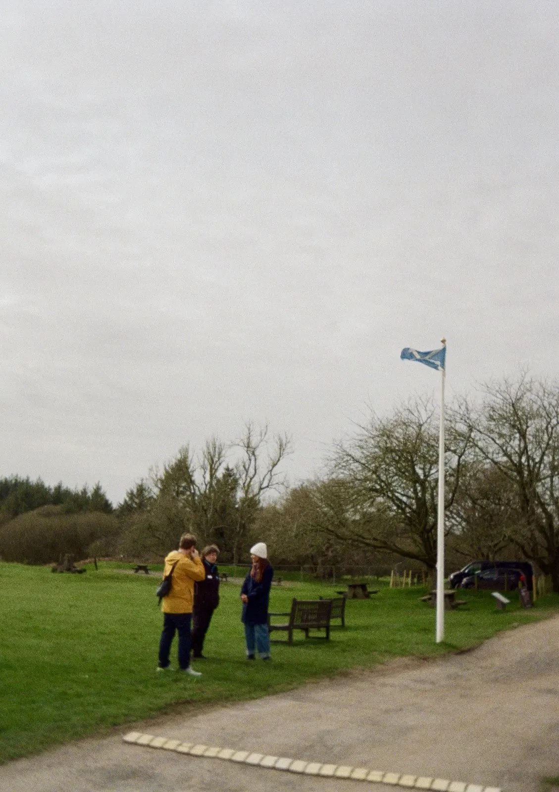 Adam Stoner, Sarah MacGillivray and Zoe Samuels-Lyons from Historic Scotland talking on a wide green lawn beside a white flagpole flying the saltire, bare trees and picnic benches behind them under a grey sky.