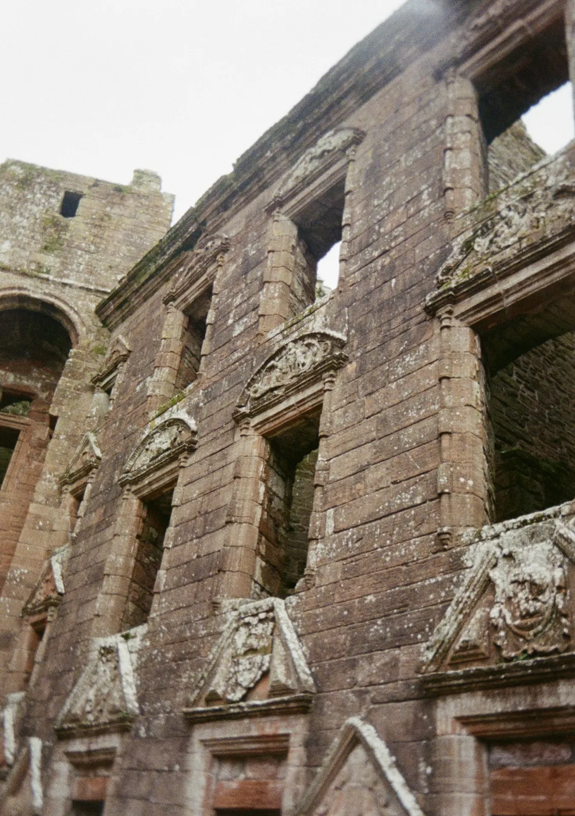 Looking up at a roofless red sandstone facade, rows of empty windows each topped with a carved stone pediment, lichen on the stonework.