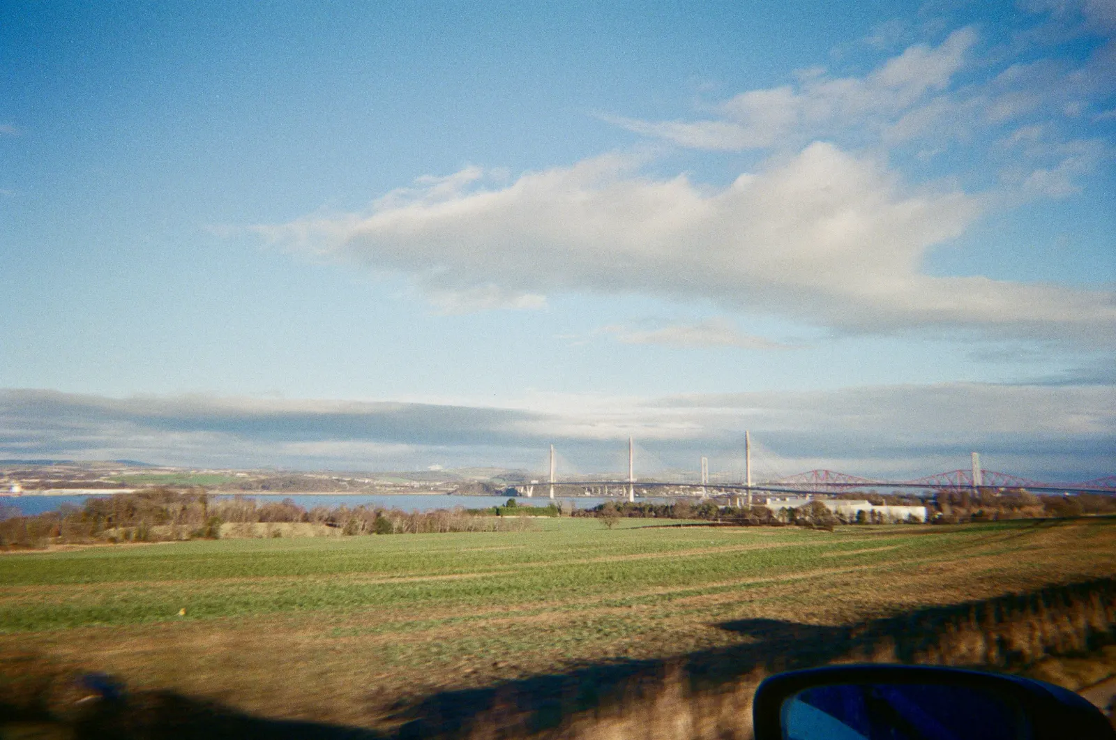 Taken from a moving car, green fields running down to the Firth of Forth, the white towers of the Queensferry Crossing and the red Forth Bridge on the horizon, a wing mirror in the corner.