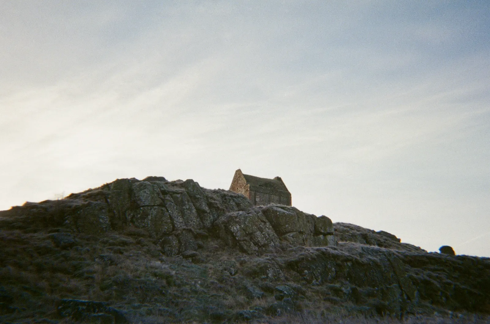 Smailholm Tower seen from below, a small stone silhouette on top of a dark rocky outcrop against a pale streaked sky.