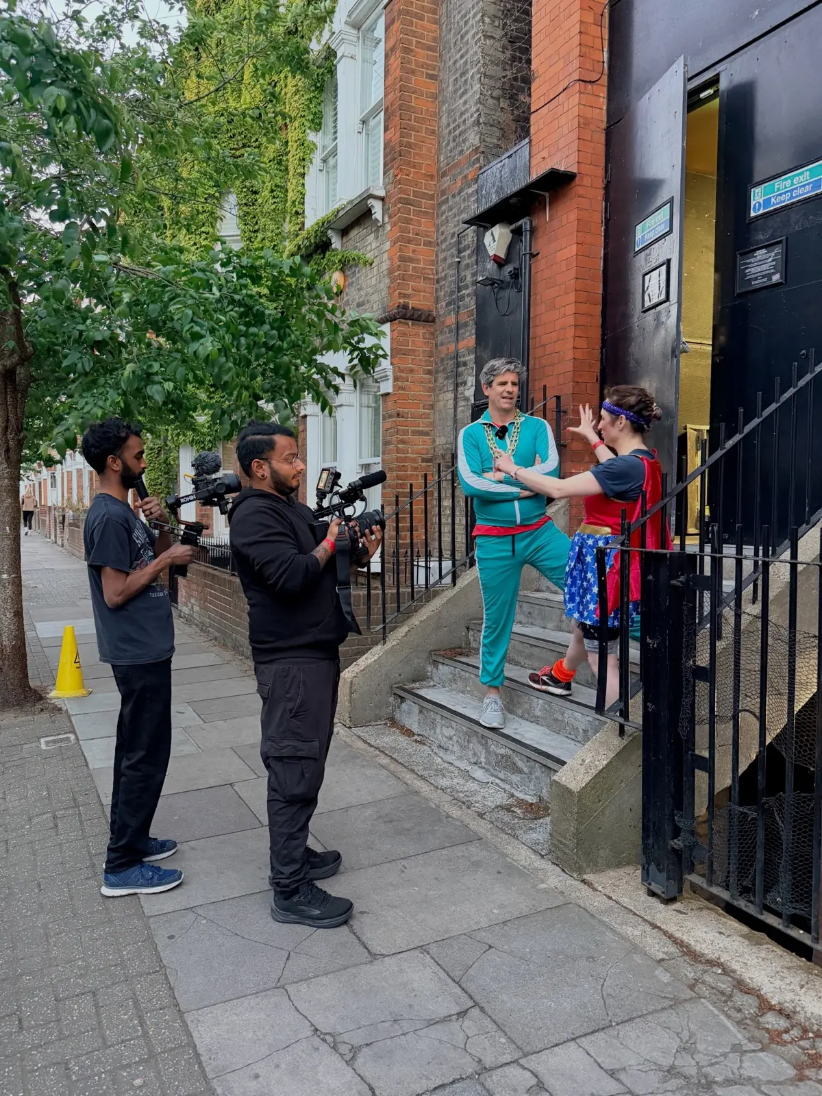 Two crew filming on a tree-lined street: a man in a turquoise tracksuit and gold chains leans on the railings of a terraced house while a wrestler in a Wonder Woman costume gestures at him from the steps.