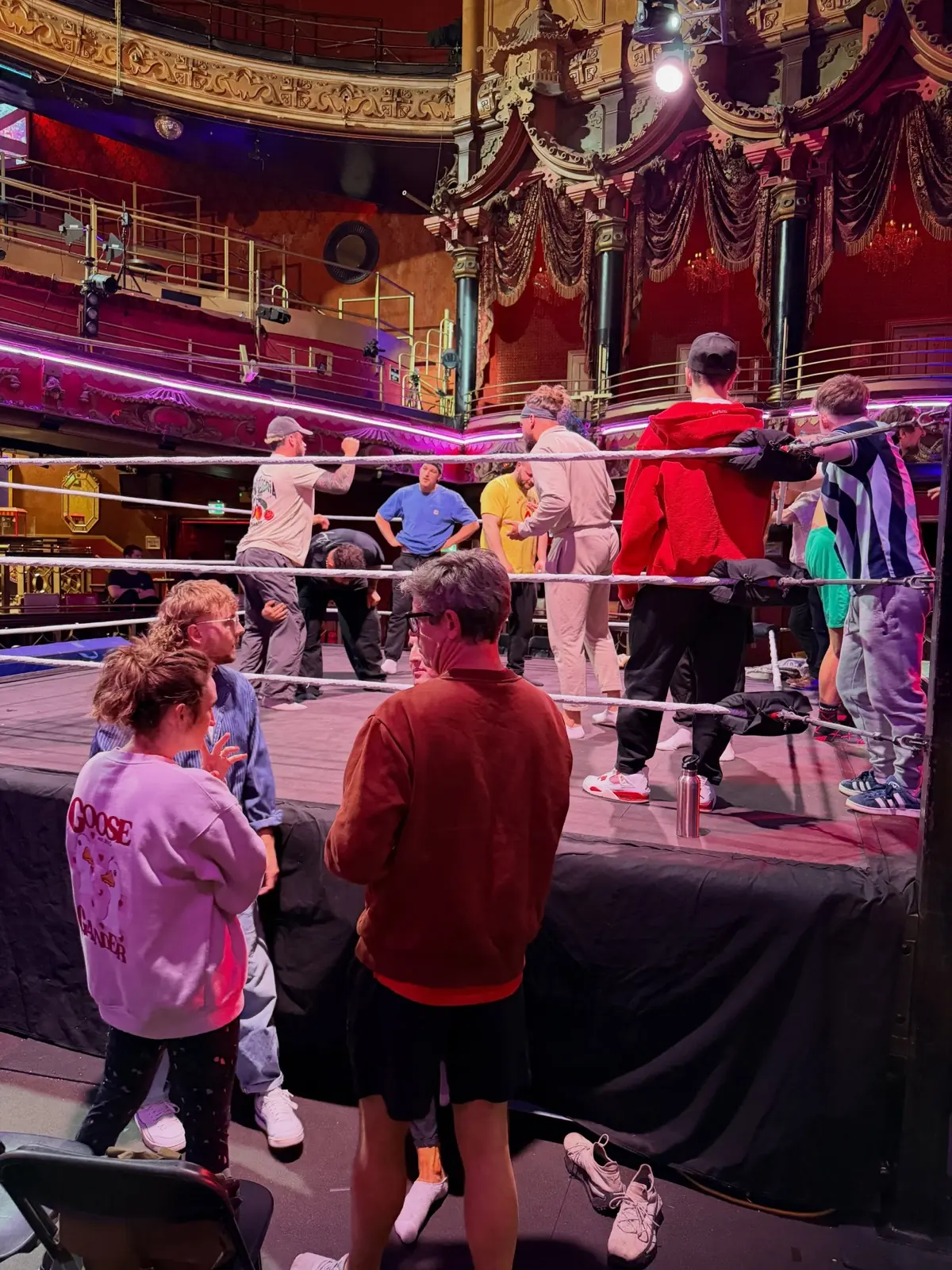 Crew and wrestlers gathered around a ring set up on the floor of an ornate old theatre, gilt balconies rising behind them.