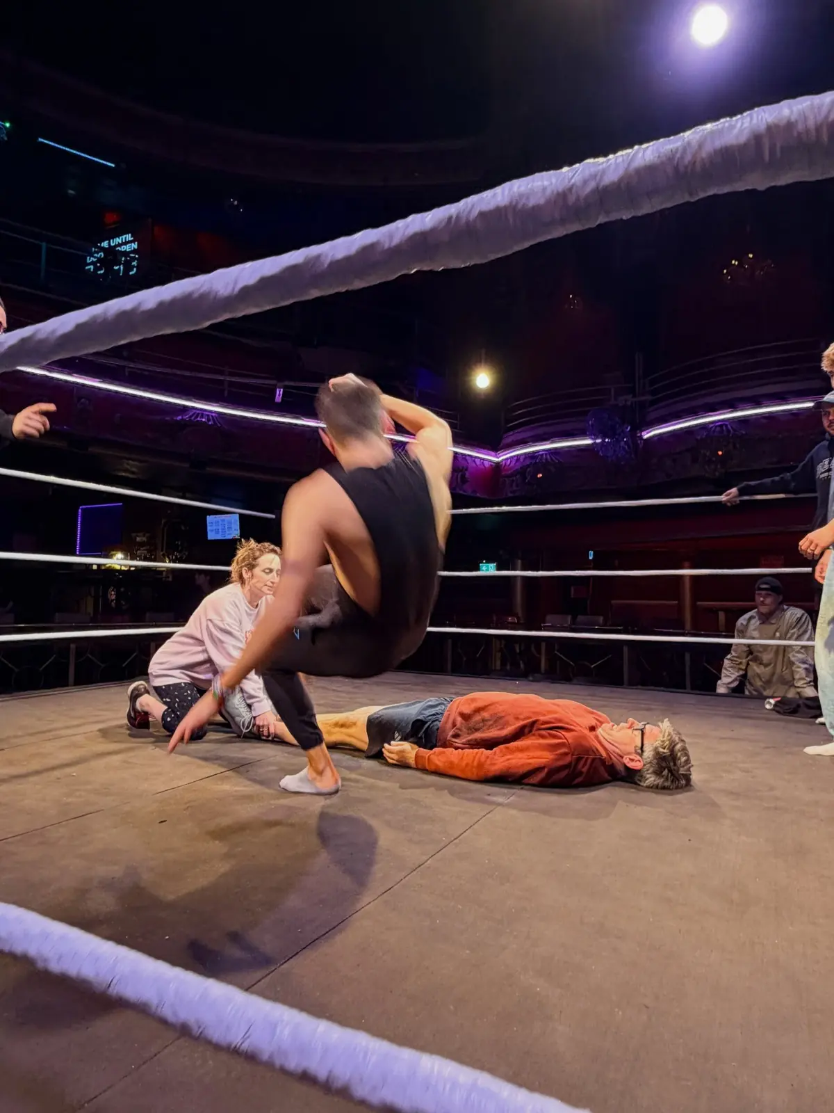 A wrestler mid-move, arched over an opponent lying flat on the mat, in the same ornate theatre.