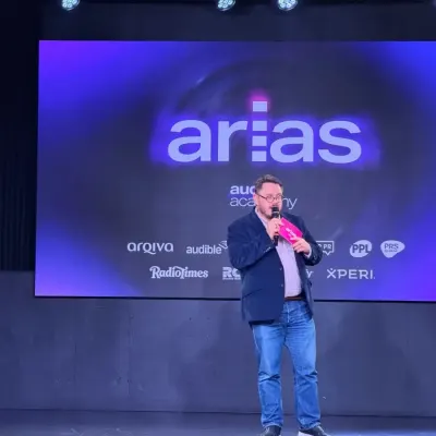 A man on stage at the ARIAS, speaking into a pink microphone in front of a large screen showing the arias and Audio Academy logos.