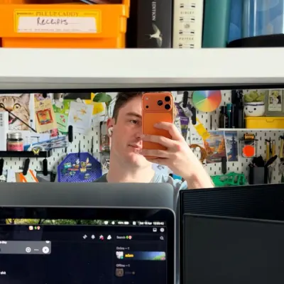 A mirror selfie at a desk, taken over two monitors. Behind, a pegboard covered in postcards, tools and an orange Penco caddy labelled 'Receipts'.