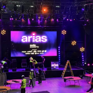 The Roundhouse main space in purple light during the afternoon setup, crew in hi-vis carrying equipment between bare round tables in front of the stage.