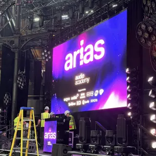 Crew in hard hats and hi-vis building the ARIAS stage under a lighting rig, a yellow stepladder in front of a big purple screen showing the arias and Audio Academy logos.