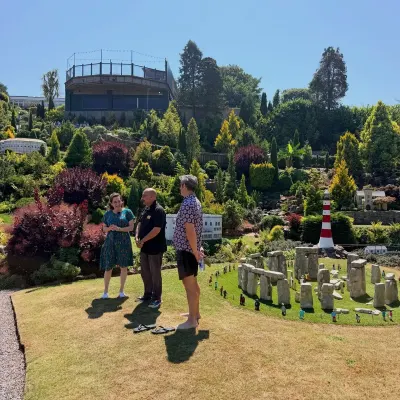 Three people standing on the lawn beside a miniature Stonehenge at Babbacombe Model Village, a red and white lighthouse and tiny buildings on the hillside behind.