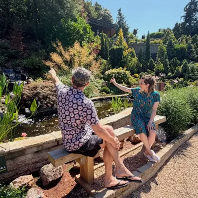 Isy Suttie and Dan Skinner on a bench beside a lily pond at Babbacombe Model Village, both pointing off at something in the miniature landscape.