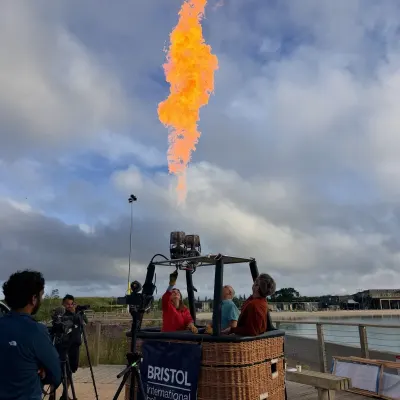 A burner flame roaring above a wicker basket stencilled Bristol International Balloon Fiesta, a camera operator filming from beside it.
