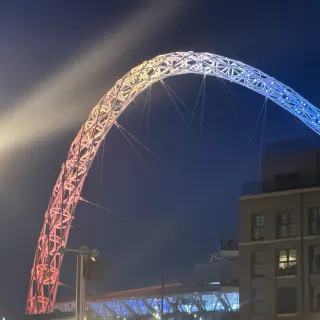 Wembley's arch lit pink fading to blue against the night sky, rising behind a block of flats and a glowing lamp post.