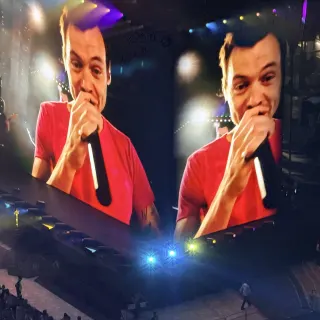 Harry Styles in a red T-shirt grinning into a microphone, shown twice across the giant stadium screens, stage lights flaring below.