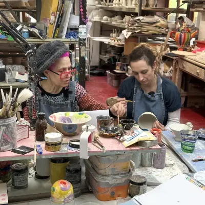 Mary Rose-Young, in pink hair and red glasses, painting pottery alongside Isy Suttie in a studio crowded with brightly coloured glazes and finished pieces.