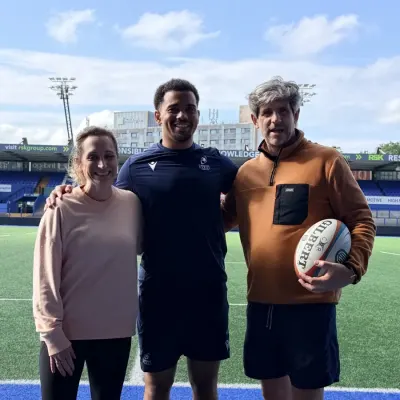 Three people arm in arm on the pitch at Cardiff Arms Park, one in Cardiff Rugby kit holding a Gilbert rugby ball, under a bright blue sky.