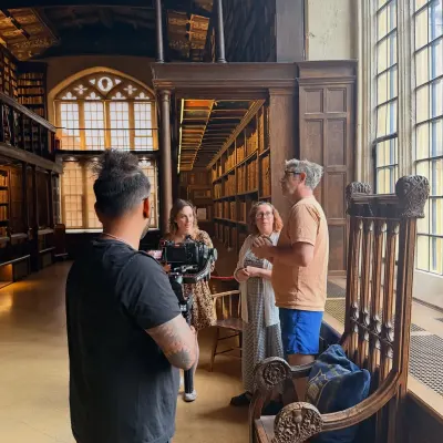A camera operator on a gimbal filming an interview between the galleried bookshelves of an Oxford library.
