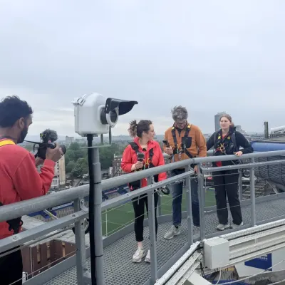 Recording on the roof walkway at Principality Stadium: two presenters in climbing harnesses, a camera operator, the pitch far below.