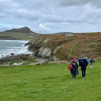 A line of people in coats and dryrobes walking a clifftop path down towards the sea in Pembrokeshire, under a pale grey sky.