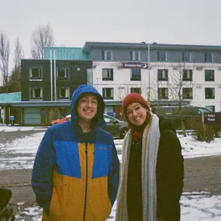 Adam Stoner in a blue and yellow hooded jacket and Sarah MacGillivray in a red beanie and long grey scarf, grinning in a snowy car park outside a Premier Inn.