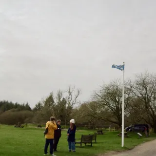 Adam Stoner, Sarah MacGillivray and Zoe Samuels-Lyons from Historic Scotland talking on a wide green lawn beside a white flagpole flying the saltire, bare trees and picnic benches behind them under a grey sky.