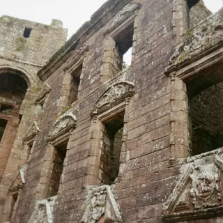 Looking up at a roofless red sandstone facade, rows of empty windows each topped with a carved stone pediment, lichen on the stonework.