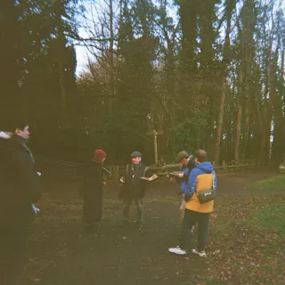 Adam Stoner, Sarah MacGillivray, Mark Watson from Historic Scotland and Ian Sheerer from Kinneil House gathered on a path among trees at Kinneil House, one of them in a flat cap reading from an open book, the sandstone wall of the house at the right edge.