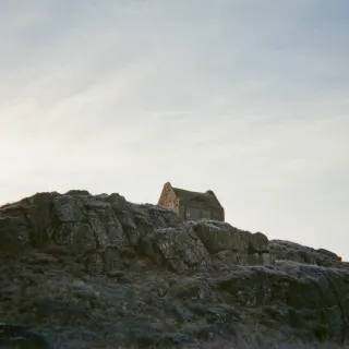Smailholm Tower seen from below, a small stone silhouette on top of a dark rocky outcrop against a pale streaked sky.
