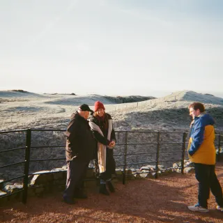 Sarah MacGillivray, Julia Hasse, Adam Stoner and an interviewee at black railings on a red gravel path, rolling hills behind them white with frost.