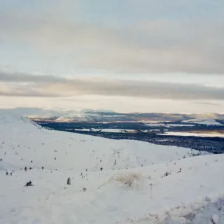 A snowy slope looking out over a dark forested valley towards distant white hills under long bands of cloud.