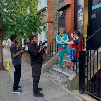 Two crew filming on a tree-lined street: a man in a turquoise tracksuit and gold chains leans on the railings of a terraced house while a wrestler in a Wonder Woman costume gestures at him from the steps.