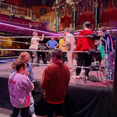 Crew and wrestlers gathered around a ring set up on the floor of an ornate old theatre, gilt balconies rising behind them.