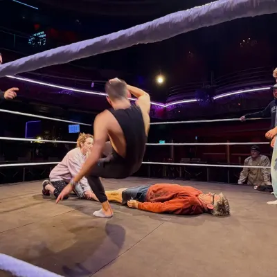 A wrestler mid-move, arched over an opponent lying flat on the mat, in the same ornate theatre.