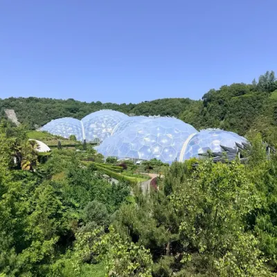 The Eden Project's biomes from across the valley, geodesic domes among Cornish greenery under a clear blue sky.
