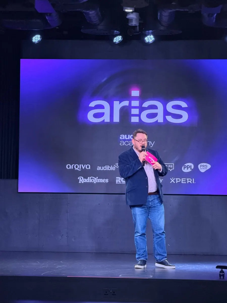A man on stage at the ARIAS, speaking into a pink microphone in front of a large screen showing the arias and Audio Academy logos.