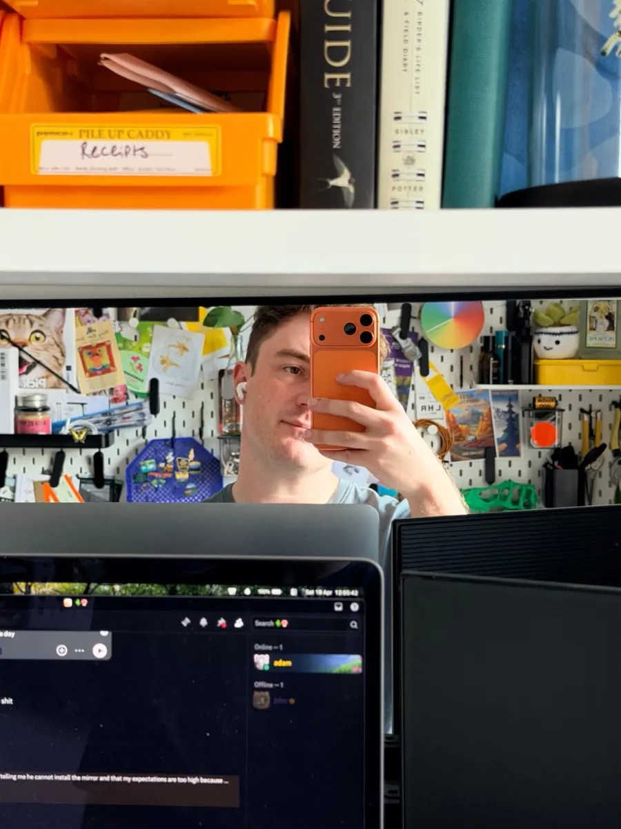 A mirror selfie at a desk, taken over two monitors. Behind, a pegboard covered in postcards, tools and an orange Penco caddy labelled 'Receipts'.