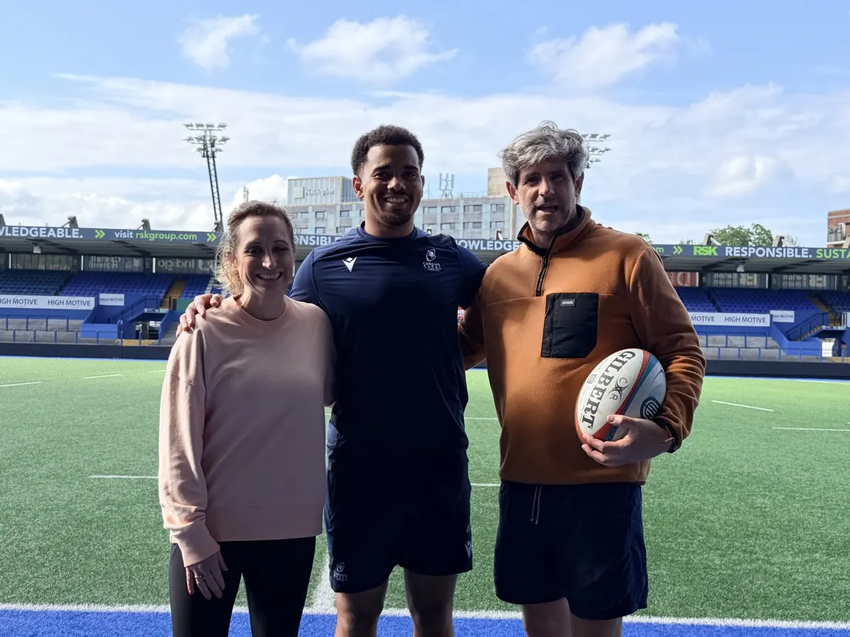 Three people arm in arm on the pitch at Cardiff Arms Park, one in Cardiff Rugby kit holding a Gilbert rugby ball, under a bright blue sky.