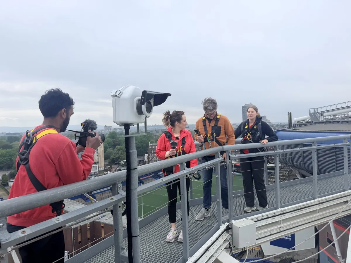 Recording on the roof walkway at Principality Stadium: two presenters in climbing harnesses, a camera operator, the pitch far below.
