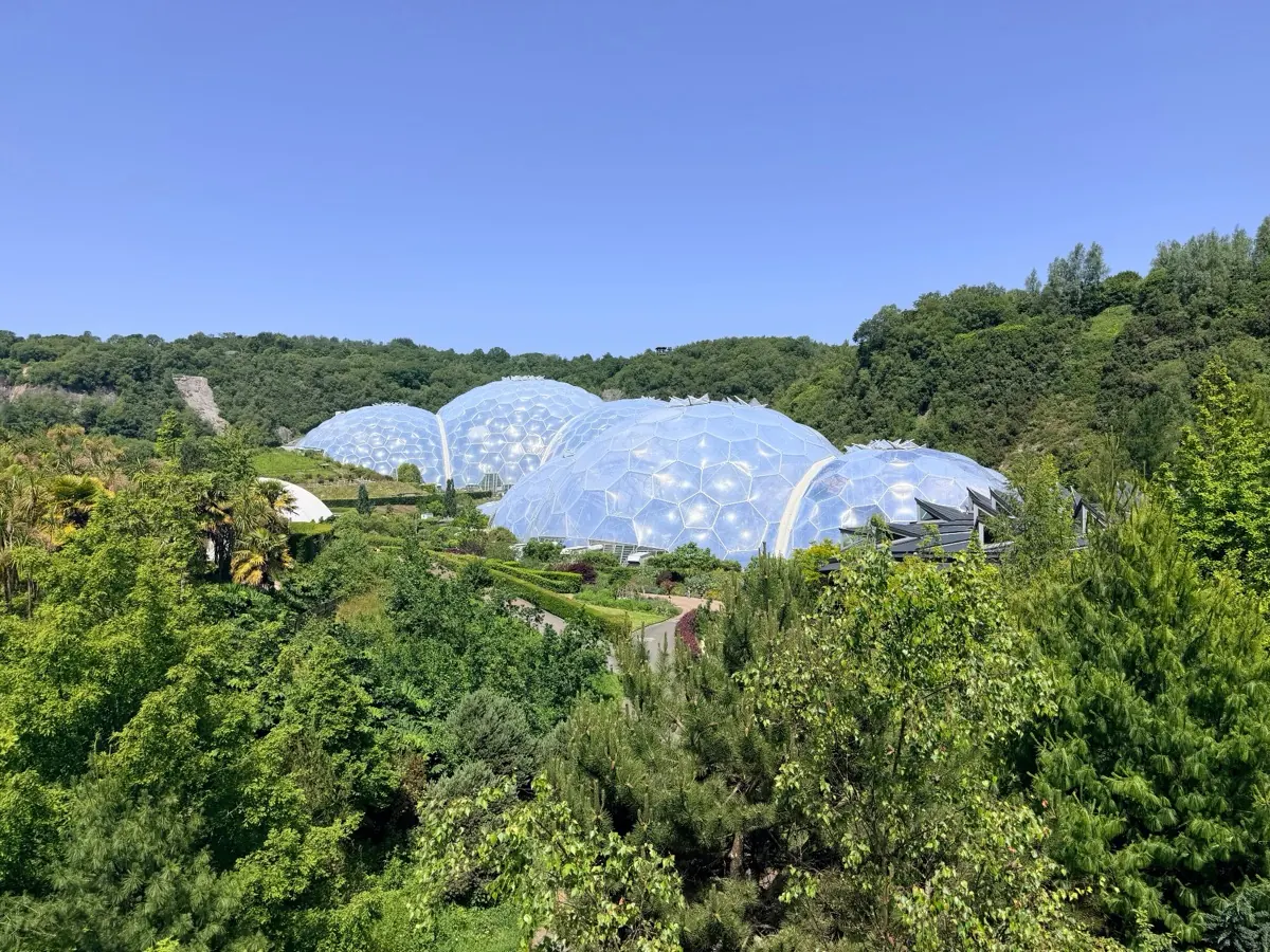The Eden Project's biomes from across the valley, geodesic domes among Cornish greenery under a clear blue sky.